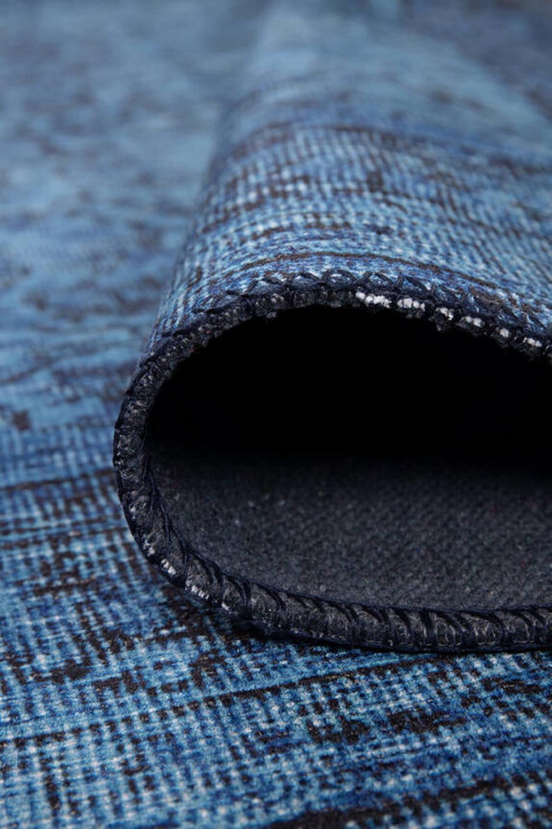 May include: Close-up of a rolled-up blue rug with a textured pattern. The rug's edge reveals a dark blue backing and a stitched border. The rug's surface has a faded, distressed appearance, with shades of blue and black.