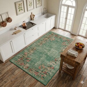 May include: A rectangular, distressed-look rug with a faded green background and a floral border in shades of pink and peach. The rug is placed in a kitchen setting with white cabinets and wooden floors. The rug's dimensions are approximately 5' x 8'.