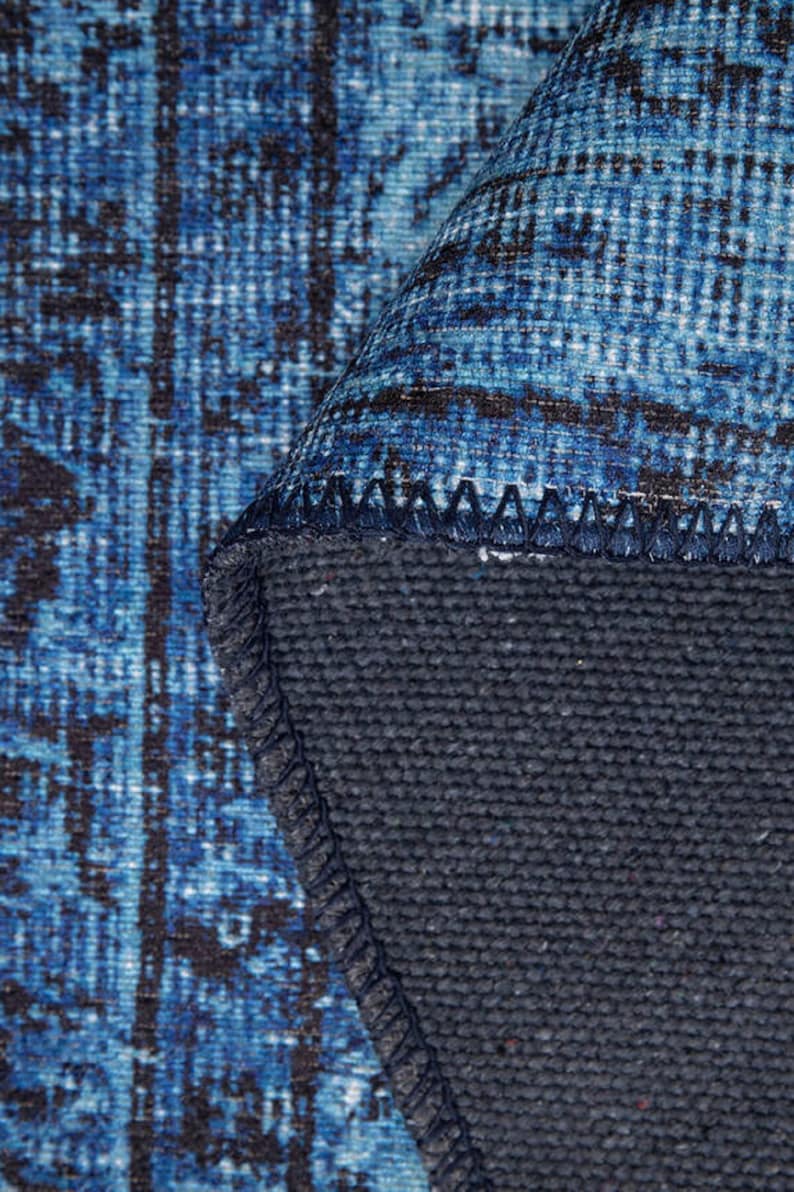 May include: Close-up of a rolled-up blue and black patterned rug. The rug's design features a distressed, vintage aesthetic with a dark blue backing. The edges are neatly stitched, showcasing the rug's construction.