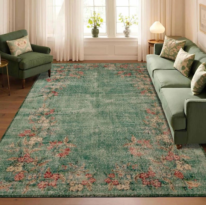 May include: A large, rectangular, distressed-look rug with a teal background and a floral border in shades of pink, cream, and green. The rug is placed in a living room setting with green furniture and natural light.