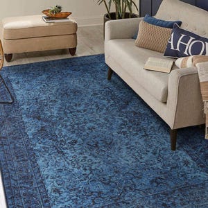 May include: A blue area rug with a distressed, floral pattern. The rug has a dark blue border and a lighter blue center. The rug is placed in a living room setting with a sofa, ottoman, and plants.