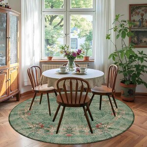 May include: A dining room scene featuring a round, green floral rug beneath a wooden table and four matching chairs. The table is set with a lace tablecloth, teacups, and a vase of flowers. A large window and a potted plant are also visible.