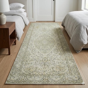 Puede incluir: Una alfombra rectangular color crema con un intrincado diseño floral descolorido. La alfombra presenta un diseño de medallón central y un borde detallado en tonos verdes y grises. Se coloca entre dos camas en un dormitorio.