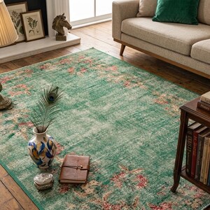 May include: A distressed green area rug with a floral pattern in pink and cream tones. The rug is placed in a living room setting, with a sofa, side table, and a decorative lamp. A peacock feather is in a vase.