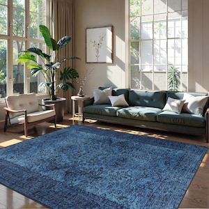 May include: A large, rectangular blue rug with a detailed floral pattern dominates the foreground of a living room scene. The rug is framed by a darker border. A teal sofa with white pillows, a wooden chair, and a large potted plant are also visible.