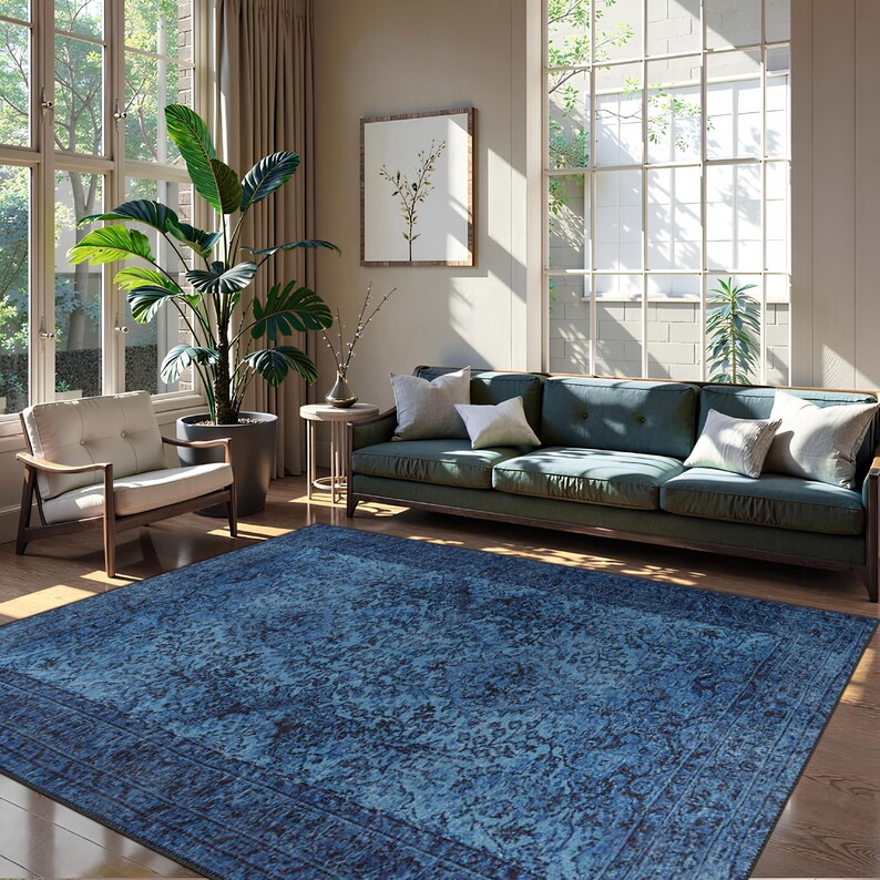 May include: A large, rectangular blue rug with a detailed floral pattern dominates the foreground of a living room scene. The rug is framed by a darker border. A teal sofa with white pillows, a wooden chair, and a large potted plant are also visible.