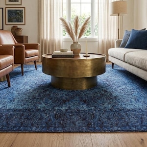 May include: A living room featuring a large blue area rug with a distressed pattern. The room includes brown leather armchairs, a beige sofa with decorative pillows, and a round brass coffee table. The room is well-lit with natural light.