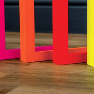 May include: Five L-shaped objects arranged in a row, each featuring a vibrant neon finish. The colours from left to right are hot pink, orange, red, yellow, and lime green. Each piece has a flat base and a vertical upright section.