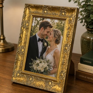 Op de afbeelding: Een rechthoekige fotolijst met een sierlijke, verweerde gouden afwerking en verhoogde decoratieve krulpatronen. De lijst bevat een foto van een koppel in trouwkleding, waarbij de persoon links een zwart pak draagt en de persoon rechts een witte kanten jurk met een boeket witte rozen.