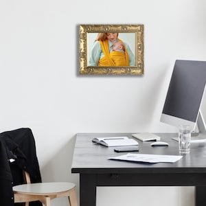 Op de afbeelding: Een rechthoekige fotolijst met een sierlijke, antieke gouden afwerking en ingewikkelde uitgesneden details. De lijst bevat een foto van een persoon die een felgele stoffen babydrager draagt en een kleine baby dicht tegen de borst houdt.