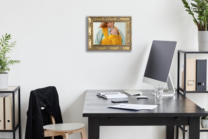 Op de afbeelding: Een rechthoekige fotolijst met een sierlijke, antieke gouden afwerking en ingewikkelde uitgesneden details. De lijst bevat een foto van een persoon die een felgele stoffen babydrager draagt en een kleine baby dicht tegen de borst houdt.