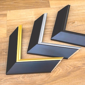 May include: Three black picture frame corners with different interior colors: gold, silver, and black. The frames are angled and have a beveled edge. The background is a light brown wooden surface.