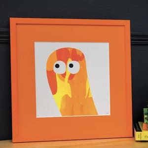 May include: A square framed print featuring an abstract orange and yellow creature with two large white eyes and black pupils. The artwork is encased in a bright orange wooden frame, creating a bold contrast against the white background of the print.