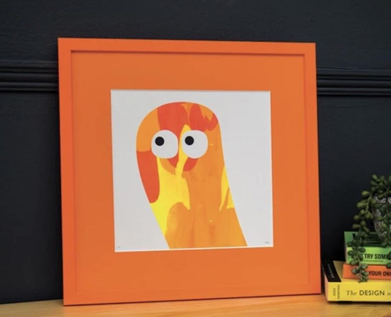 May include: A square framed print featuring an abstract orange and yellow creature with two large white eyes and black pupils. The artwork is encased in a bright orange wooden frame, creating a bold contrast against the white background of the print.