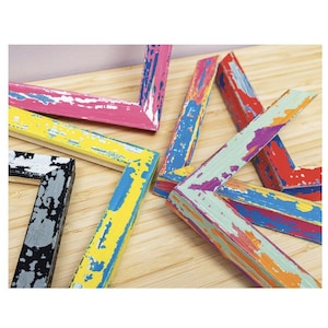 May include: A selection of distressed wooden picture frame corners in various colours. The frames are painted in pink, yellow, blue, red, and green, with a weathered, peeling paint effect. The frames are arranged on a light wood surface.