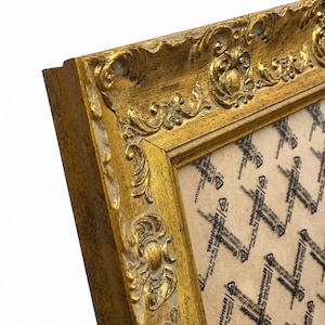 Op de afbeelding: Een rechthoekige fotolijst met een sierlijk, verhoogd goudkleurig reli&euml;fpatroon langs de randen. De lijst heeft een verweerde afwerking met zichtbare witte basislagen. Een beschermvel met herhalende zwarte tekst en symbolen bedekt het midden.