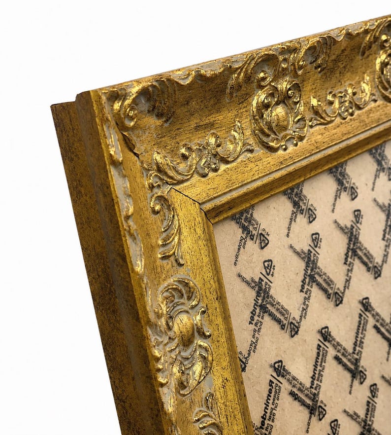 Op de afbeelding: Een rechthoekige fotolijst met een sierlijk, verhoogd goudkleurig reli&euml;fpatroon langs de randen. De lijst heeft een verweerde afwerking met zichtbare witte basislagen. Een beschermvel met herhalende zwarte tekst en symbolen bedekt het midden.