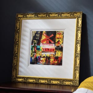 Op de afbeelding: Een ingelijste kunstdruk van de Moulin Rouge in Parijs, met een levendige rode windmolen en een straatsc&egrave;ne in gele en zwarte tinten. Het kunstwerk zit in een sierlijke, getextureerde gouden lijst met een brede witte passe-partout rand.