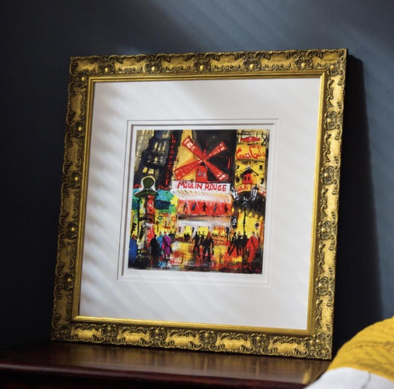 Op de afbeelding: Een ingelijste kunstdruk van de Moulin Rouge in Parijs, met een levendige rode windmolen en een straatsc&egrave;ne in gele en zwarte tinten. Het kunstwerk zit in een sierlijke, getextureerde gouden lijst met een brede witte passe-partout rand.