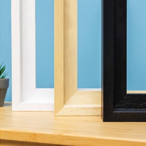 Puede incluir: Se muestran tres marcos de fotos, uno blanco, uno de madera natural y uno negro, uno al lado del otro. Los marcos son todos del mismo tamaño y forma, y están todos de pie sobre una superficie de madera.