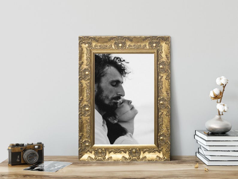Op de afbeelding: Een rechthoekige fotolijst met een sierlijke, reli&euml;f gouden afwerking met een zwart-witfoto van twee mensen die tegen elkaar aan leunen. De lijst is voorzien van ingewikkeld krulwerk en verhoogde cirkelvormige details langs de rand.