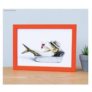 May include: A rectangular orange frame containing a print of a sardine wearing a straw hat, resting inside a metal tin, and holding a small cocktail glass with a straw. The artwork features the text Artwork by Wolf & Blue in the top corner.
