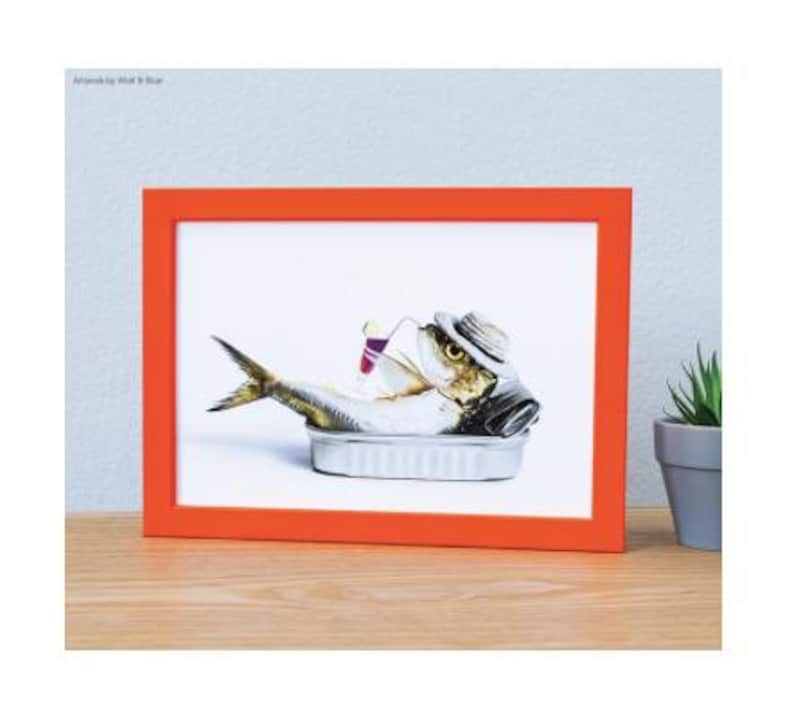 May include: A rectangular orange frame containing a print of a sardine wearing a straw hat, resting inside a metal tin, and holding a small cocktail glass with a straw. The artwork features the text Artwork by Wolf & Blue in the top corner.
