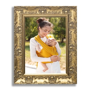 Op de afbeelding: Een rechthoekige fotolijst met een versierde, in reli&euml;f gemaakte gouden afwerking met ingewikkelde krullen en bloemmotieven. De lijst bevat een foto van een persoon die een mosterdgele stoffen babydrager draagt met een baby erin.