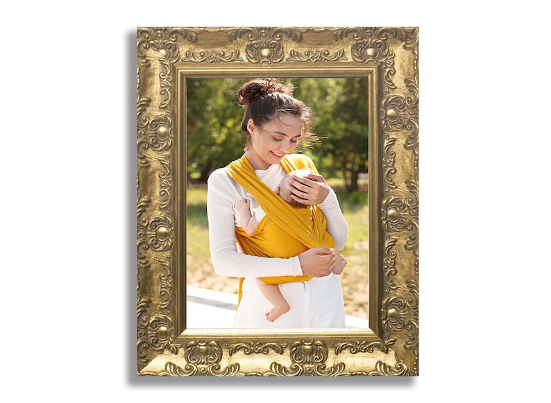 Op de afbeelding: Een rechthoekige fotolijst met een versierde, in reli&euml;f gemaakte gouden afwerking met ingewikkelde krullen en bloemmotieven. De lijst bevat een foto van een persoon die een mosterdgele stoffen babydrager draagt met een baby erin.