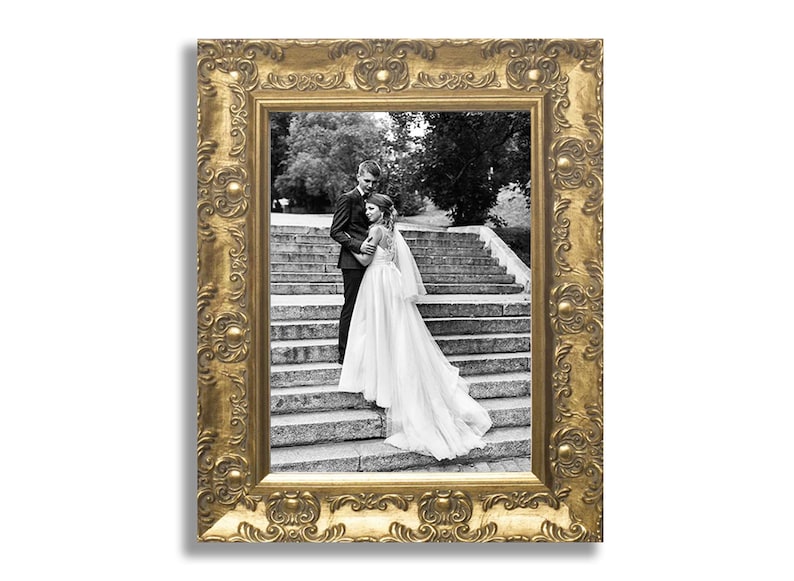 Op de afbeelding: Een rechthoekige fotolijst met een sierlijke, reli&euml;f gouden afwerking met herhalende bloem- en cirkelmotieven. De lijst bevat een zwart-witfoto van twee personen die op een stenen trap staan.