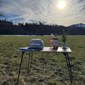 Könnte beinhalten: Ein grauer und schwarzer Klapptisch mit einer grauen Baseballkappe mit dem Wort "detowa" darauf, zwei Glasflaschen und eine kleine Topfpflanze. Der Tisch steht auf einer grasbewachsenen Wiese mit einem strahlend blauen Himmel und einer Sonne, die im Hintergrund scheint.