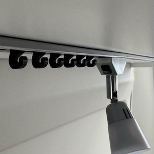 Può includere: Un sistema di binari per tende montato a soffitto con ganci neri. Un faretto argentato è attaccato, rivolto verso il basso. Lo sfondo è una parete bianca sporca. Ideale per interni moderni.