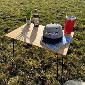 Könnte beinhalten: Ein kleiner, hellfarbiger Holztisch mit schwarzen Haarnadelbeinen. Auf dem Tisch befinden sich eine grau-schwarze Baseballkappe mit dem Wort "delta", ein roter Becher, eine kleine Topfpflanze, eine Bierflasche und eine Zeitschrift.