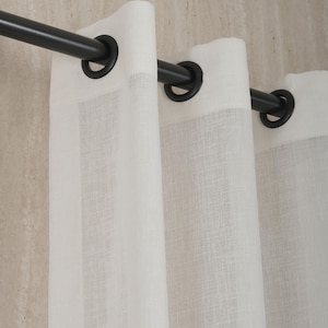 Puede incluir: Primer plano de cortinas de lino blanco colgadas de una barra de metal negro. Las cortinas tienen grandes ojales y un aspecto texturizado. El fondo es una pared neutra de color claro.