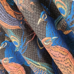 May include: A dark gray fabric featuring a repeating pattern of peacocks in vibrant shades of blue, orange, and teal. The woven material displays detailed bird figures with prominent plumage and crests, creating a textured surface with a rich color palette.
