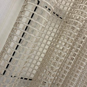 Puede incluir: Un panel de cortina beige transparente con un patrón de cuadrícula estructurado. La tela está compuesta por bandas tejidas horizontales y verticales que crean una serie de espacios rectangulares abiertos, resultando en un aspecto texturizado y semitransparente.