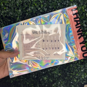 Puede incluir: Un kit de medición de uñas holográfico en una bolsa iridiscente transparente. El kit incluye una tarjeta con instrucciones y puntas de uñas numeradas para ambas manos. El texto "NAIL SIZING KIT" es visible.