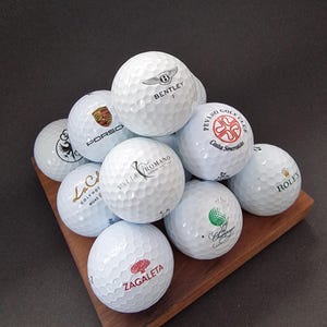 Może przedstawiać: Kolekcja białych piłek golfowych z różnymi logo, w tym Bentley, Porsche i Rolex, ułożonych na drewnianym kwadracie. Piłki golfowe są wgłębione i wyglądają na nowe. Logo są w różnych kolorach, w tym czarnym, złotym i zielonym.