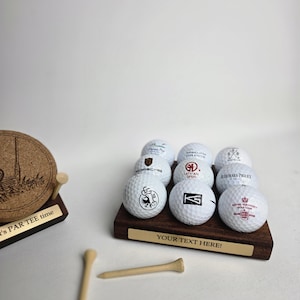 Könnte beinhalten: Eine Präsentation von Golfbällen mit verschiedenen Logos auf einem dunklen Holzsockel. Der Sockel hat ein goldfarbenes Schild mit dem Text "YOUR TEXT HERE!". Zwei Golf-Tees aus Holz befinden sich im Vordergrund.
