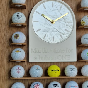 May include: A wooden golf ball display rack featuring a central arched clock with a white marble-patterned face, gold hands, and the engraved text 'Martin - time for a hole in ONE'. The rack holds numerous white and yellow golf balls on shelves.