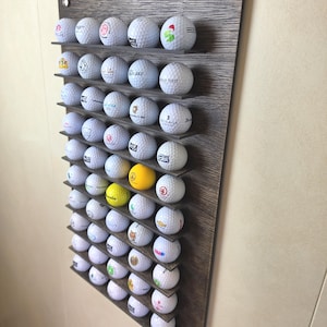 Könnte beinhalten: Ein wandmontiertes Regal aus dunklem Holz mit strukturierter Maserung, das zwölf horizontale Ablagen umfasst, auf denen eine Sammlung weißer Golfbälle sowie zwei gelbe Golfbälle mit verschiedenen Logos und Schriftzügen präsentiert werden.