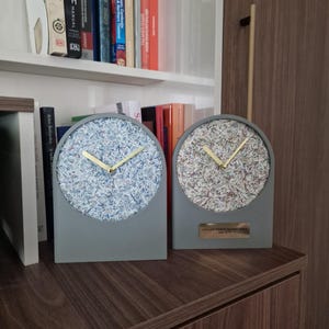 May include: Two gray, arched-top clocks with light blue and multicolored faces, gold hands, and a gray base. One clock has a small plaque. The clocks are on a dark wood surface with books in the background.
