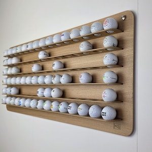 May include: A rectangular light-coloured wooden wall-mounted display rack featuring multiple horizontal shelves with circular cutouts designed to hold and organise a collection of white golf balls, with a small logo etched in the bottom corner.
