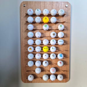 Peut inclure: Un présentoir en bois pour balles de golf avec plusieurs étagères, contenant de nombreuses balles de golf blanches. Les balles de golf présentent divers logos et motifs dans des couleurs comme le vert, le jaune et le rouge. Le présentoir est monté sur un mur clair.