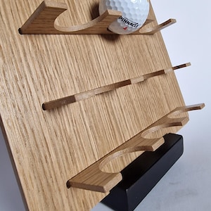 Könnte beinhalten: Ein hellbrauner Golfball-Präsentationsständer aus Holz mit drei horizontalen Ablagen und kreisförmigen Aussparungen für Golfbälle. Ein weißer Pinnacle-Golfball mit einer roten Nummer vier liegt in der obersten Ablage des Ständers, der auf einem massiven schwarzen rechteckigen Sockel steht.