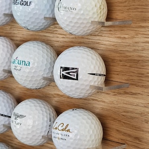 Peut inclure: Présentoir en bois avec plusieurs balles de golf blanches, chacune sur une étagère en acrylique transparente. Les balles de golf affichent des logos et du texte imprimés, dont "River Golf", "Romano", "Laguna", "Bentley" et "La Cala".