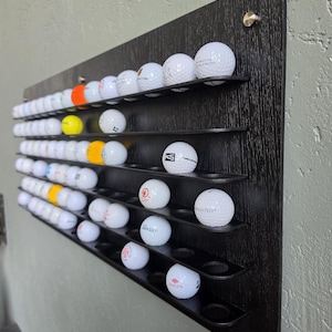 May include: A black wooden wall-mounted display rack with horizontal shelves featuring circular cutouts to hold golf balls. The rack is filled with numerous white golf balls, along with a few yellow and orange ones, arranged in rows on the dark surface.