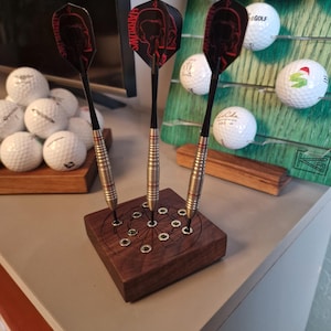 May include: Three steel-tipped darts with black shafts and black flights featuring red skull graphics, standing upright in a square dark wood display base with a circular metal-inlaid pattern and twelve holes for dart storage.