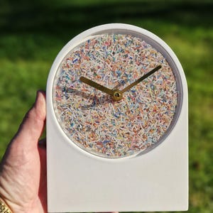 Puede incluir: Reloj de escritorio blanco con una esfera transparente llena de trozos de papel de colores. El reloj tiene manecillas doradas y un centro dorado. El reloj se sostiene en una mano con un reloj dorado.
