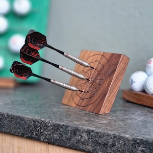 May include: Three metal darts with black and red flights featuring the Harrows logo are embedded into a rectangular wooden block engraved with a circular dartboard pattern. The darts have silver barrels with thin red rings and black shafts.